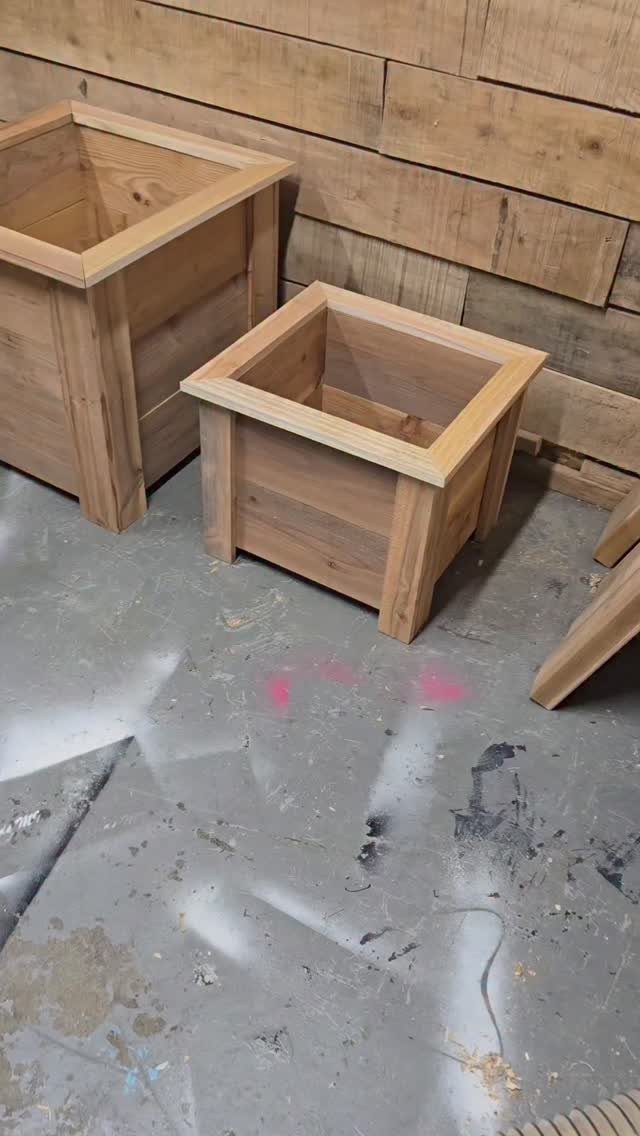 Two square wooden planter boxes sitting on a concrete floor against a wood wall.