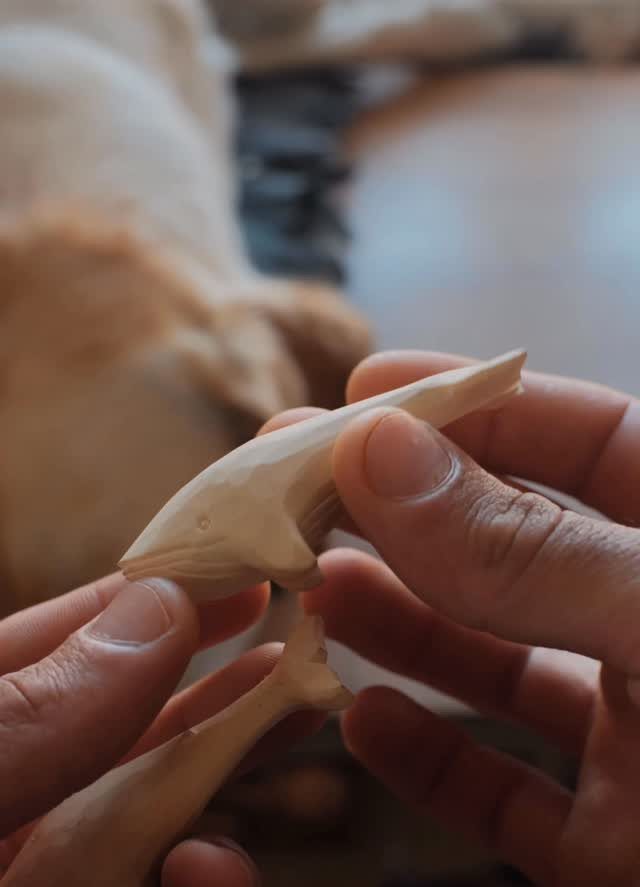 Hands holding two small, hand-carved wooden whale figures in a soft-lit setting.