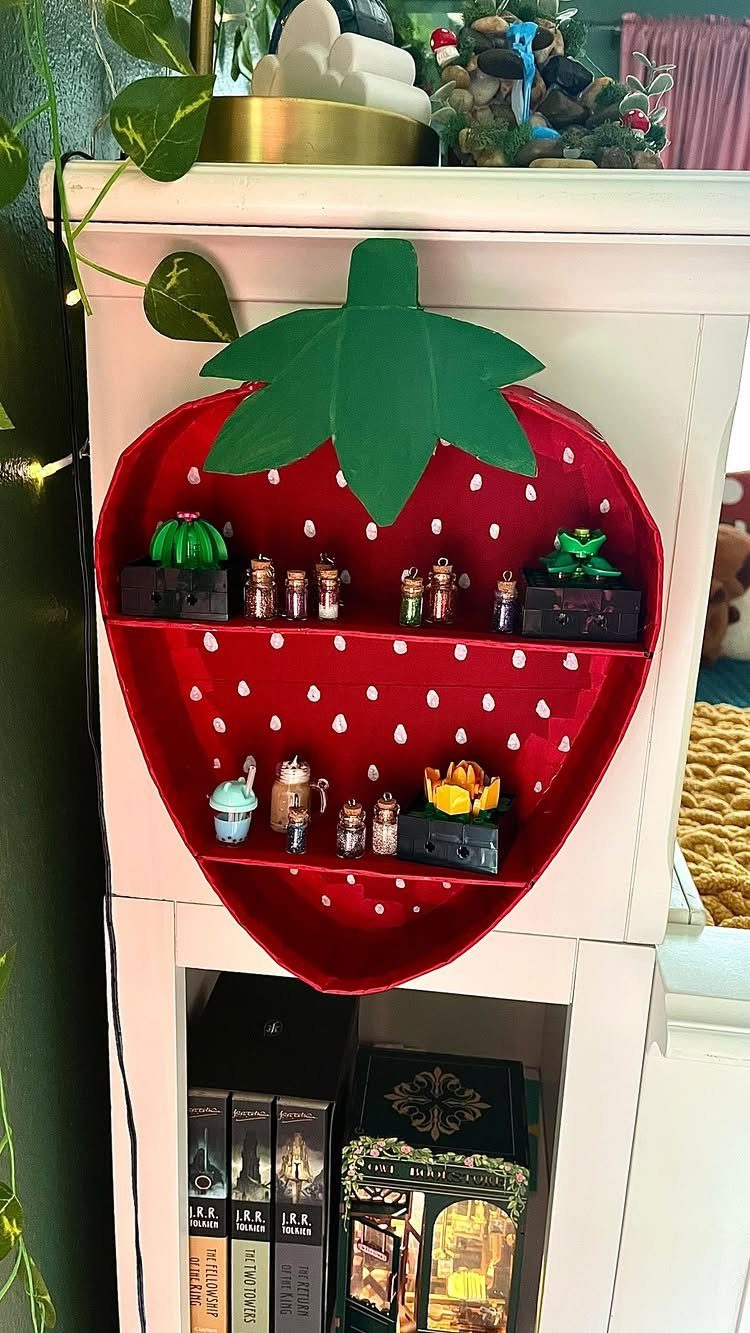 Strawberry-shaped red wall shelf holding miniature figurines and plants on a white cabinet.