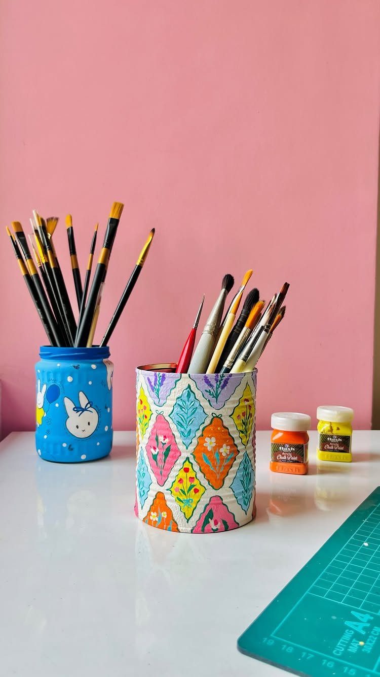 Two colorful containers holding paint brushes on a white table against pink wall.