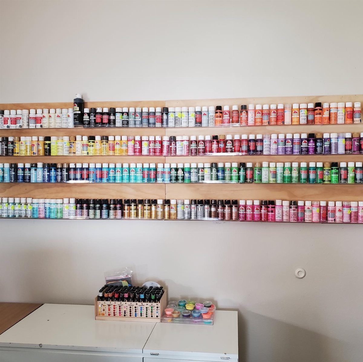 Wooden wall shelves holding organized rows of colorful paint bottles in a studio