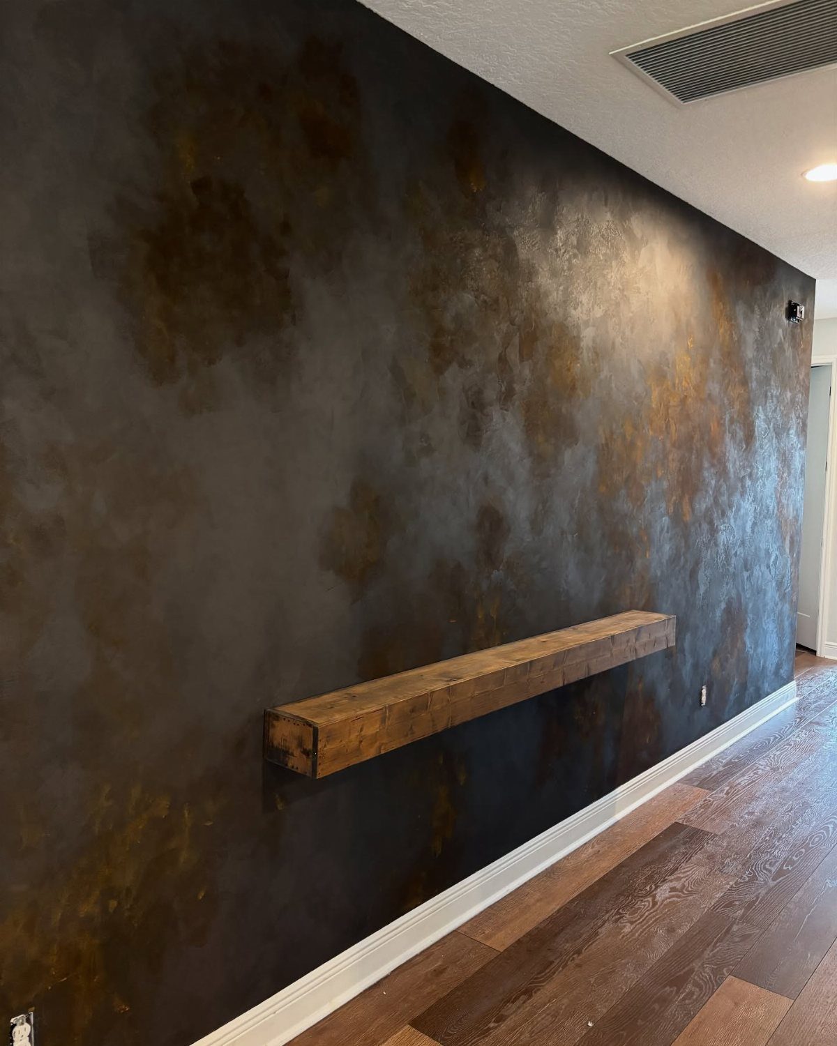 Rustic wooden floating shelf on a dark textured metallic wall with wood flooring
