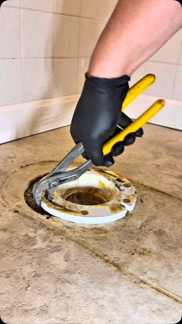 Hand in black glove using pliers to remove a toilet flange from floor