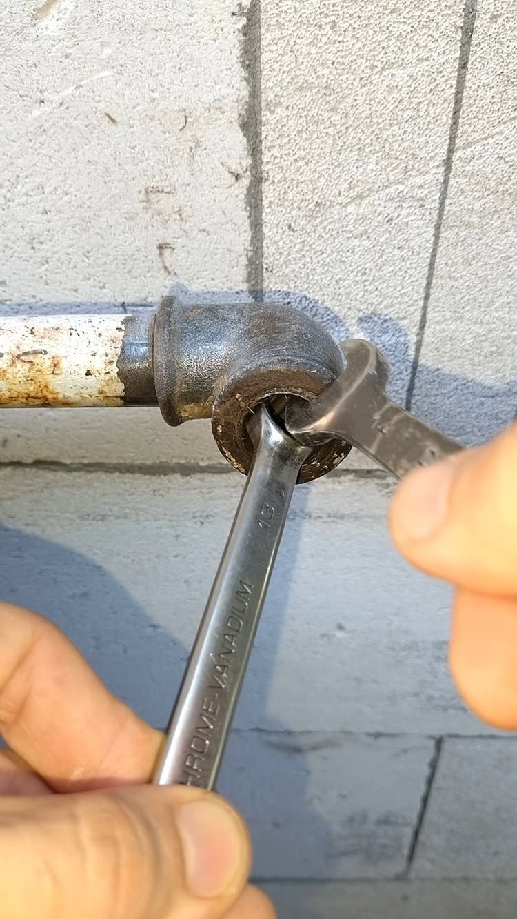 Hands using metal wrenches to repair a rusty pipe against a concrete wall.