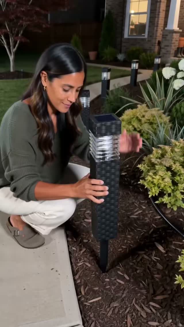 Woman installing black solar pathway lights in a garden bed along a walkway.