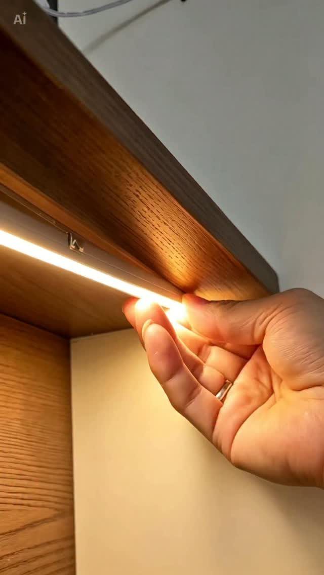 Hand installing a bright LED light strip under a brown wooden cabinet shelf.