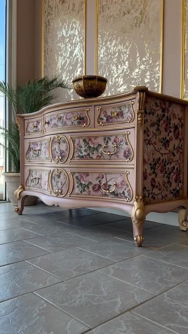 Pink vintage dresser with floral decoupage patterns and ornate gold accents on grey tiles.