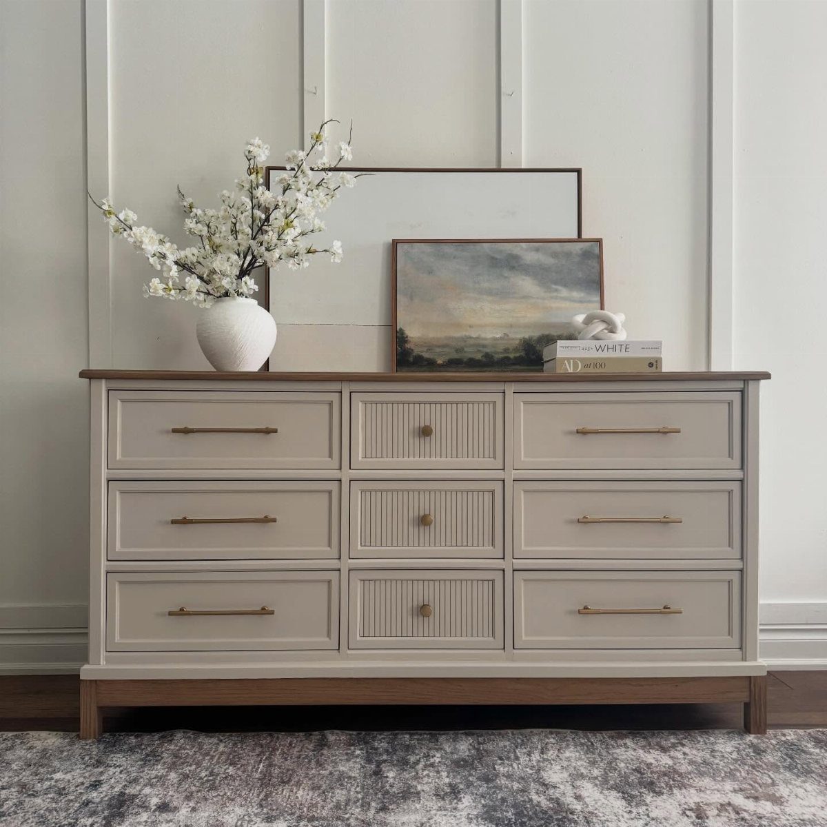 Beige dresser with gold handles and fluted drawers topped with white flowers and art.
