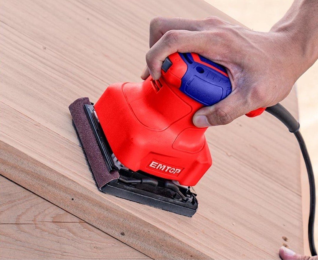 Red electric orbital sander smoothing a light brown wooden board surface.