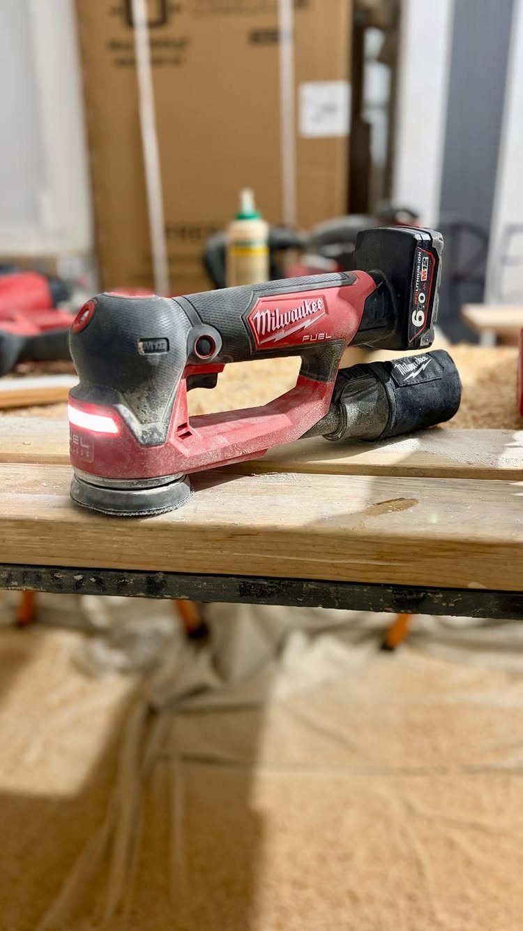 Furniture Making Tools: 11 Hand and Power Tools to Own 4 Red cordless orbital sander sitting on a light-colored wooden workbench in a workshop.
