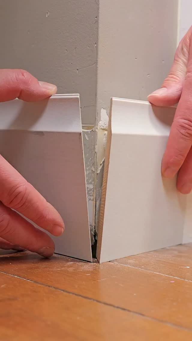 Hands fitting two white mitered baseboard pieces together in a wall corner.