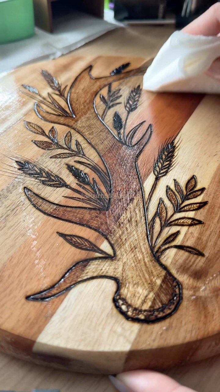 Hand applying clear finish to a wood-burned antler design on a wooden board