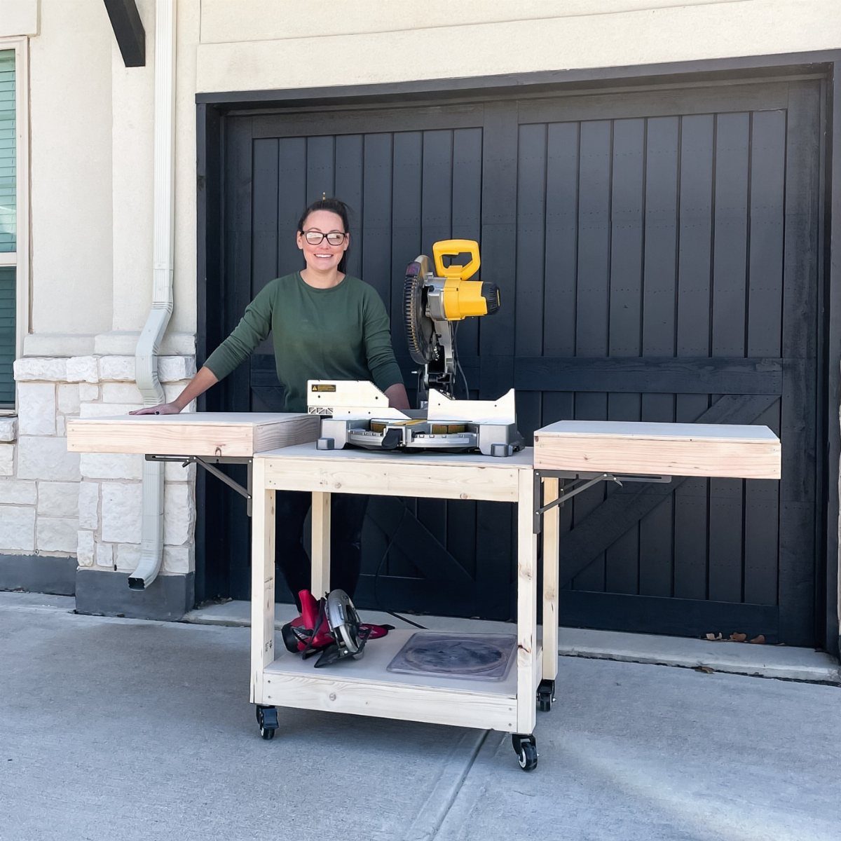 Wooden rolling miter saw stand with a lower shelf and a woman standing beside it.