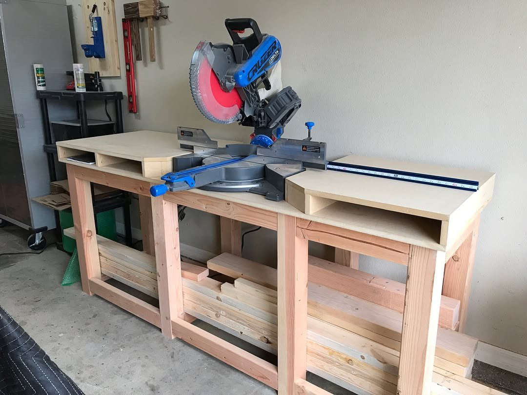 Miter saw mounted on a custom wooden workbench with lower storage shelves.