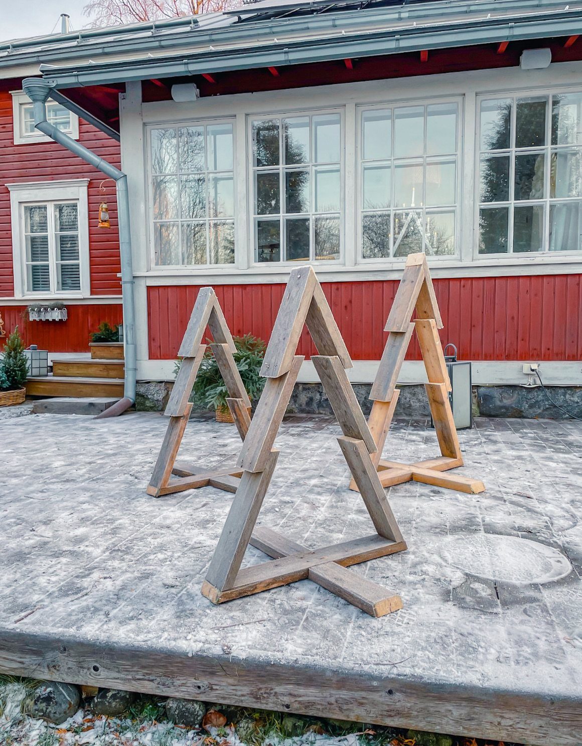 Three wooden minimalist Christmas tree structures standing on a concrete patio by a red house.
