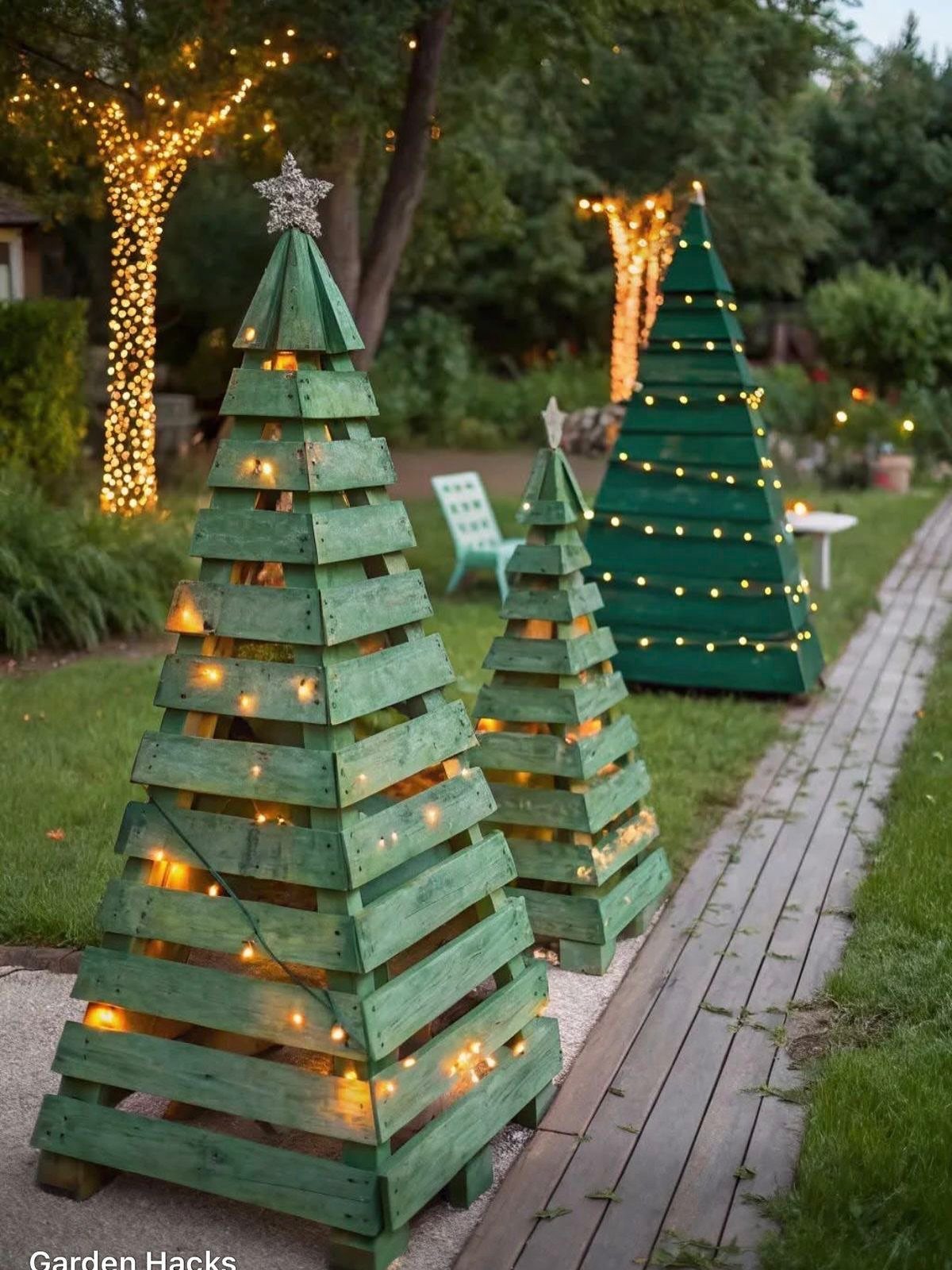 Three green wooden Christmas trees of different sizes with lights in a garden.