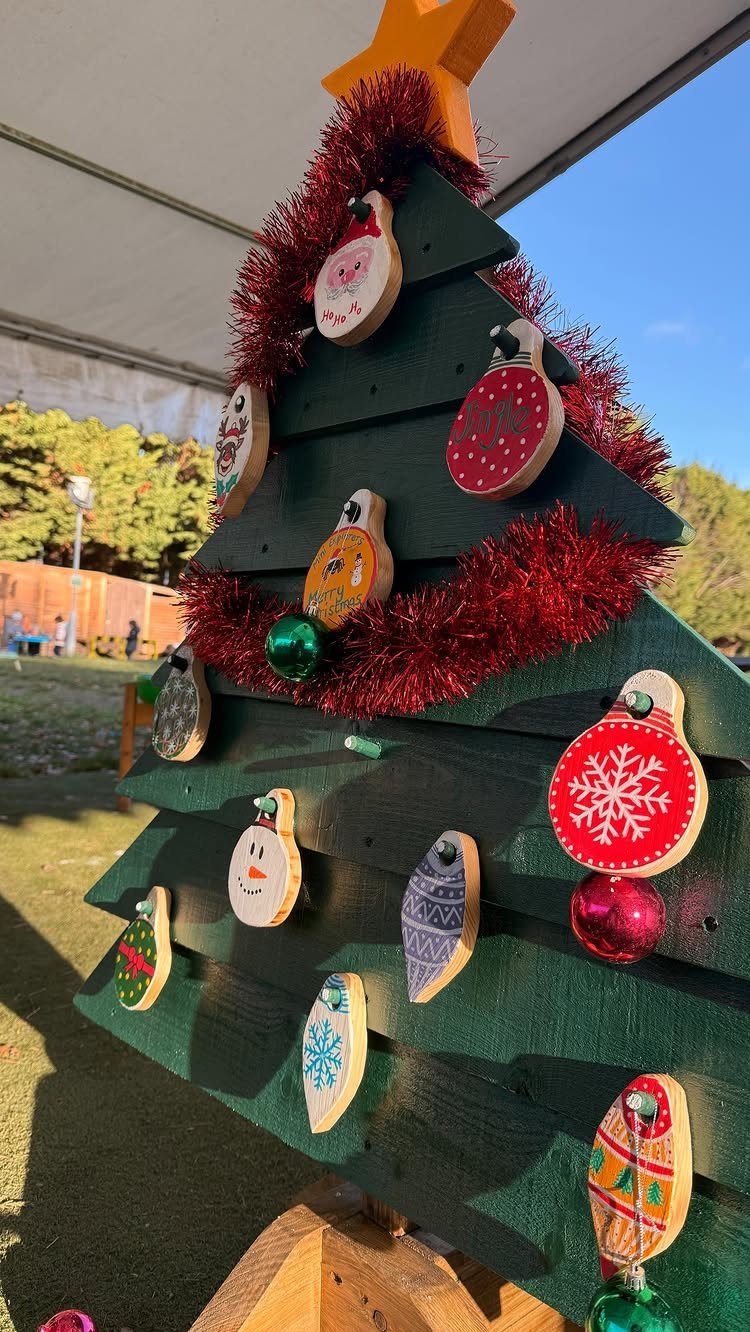 Green painted wooden Christmas tree with red tinsel and colorful wooden ornaments outdoors.