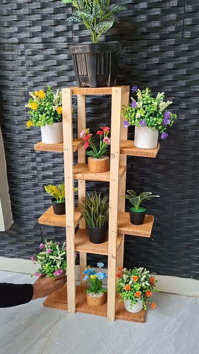 Wooden multi-tiered plant stand holding various colorful potted flowers against a textured wall
