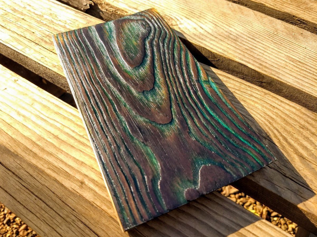 Charred wood plank with iridescent teal and purple stains on a wooden deck.