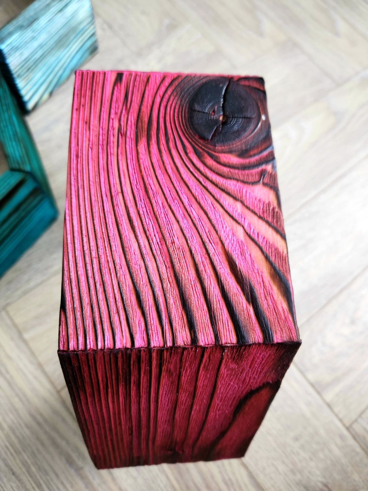 Charred wood block with deep black grain and vibrant pinkish-red stain on light flooring.