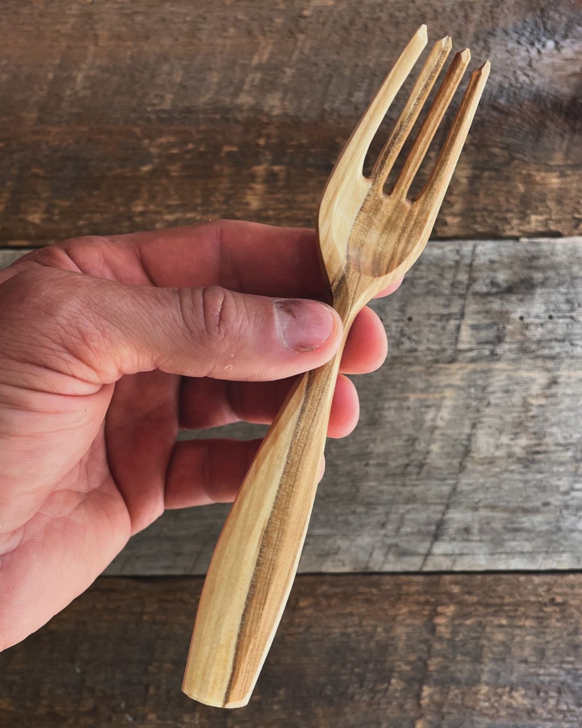 Hand-carved wooden fork held by a person against a rustic wooden backdrop