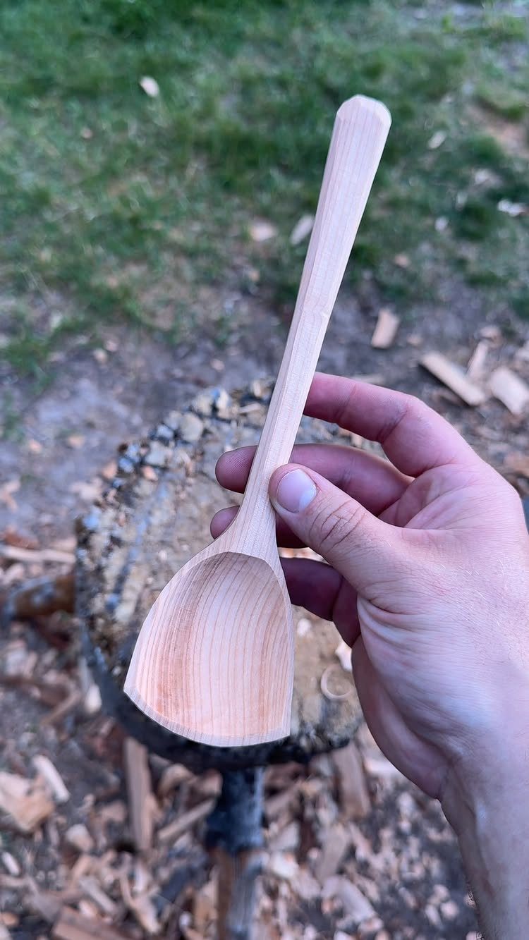 Hand-carved light wooden spoon held against a background of wood shavings and grass.