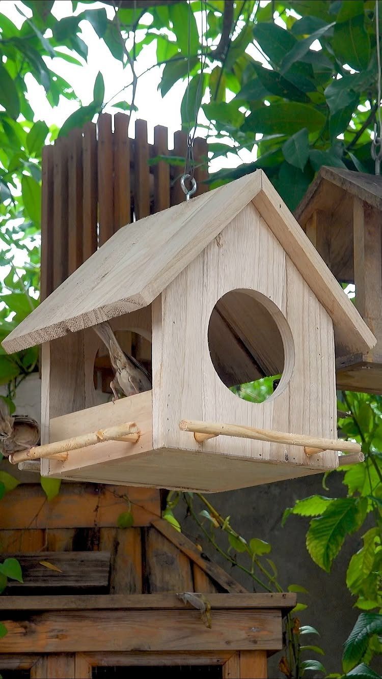 Wooden birdhouse with a peaked roof hanging among green leaves in a garden.