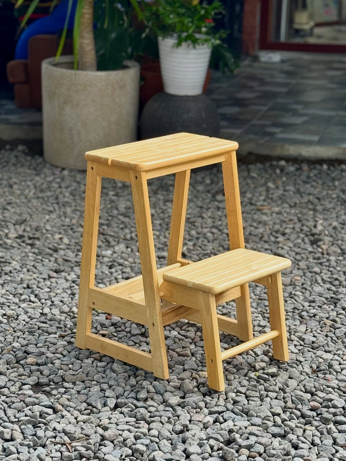 Two-step wooden stool with a natural finish standing on a grey gravel ground.