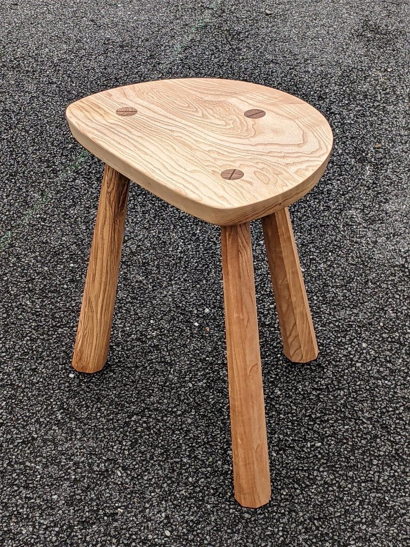 Three-legged wooden stool with an oval seat on a grey asphalt ground.