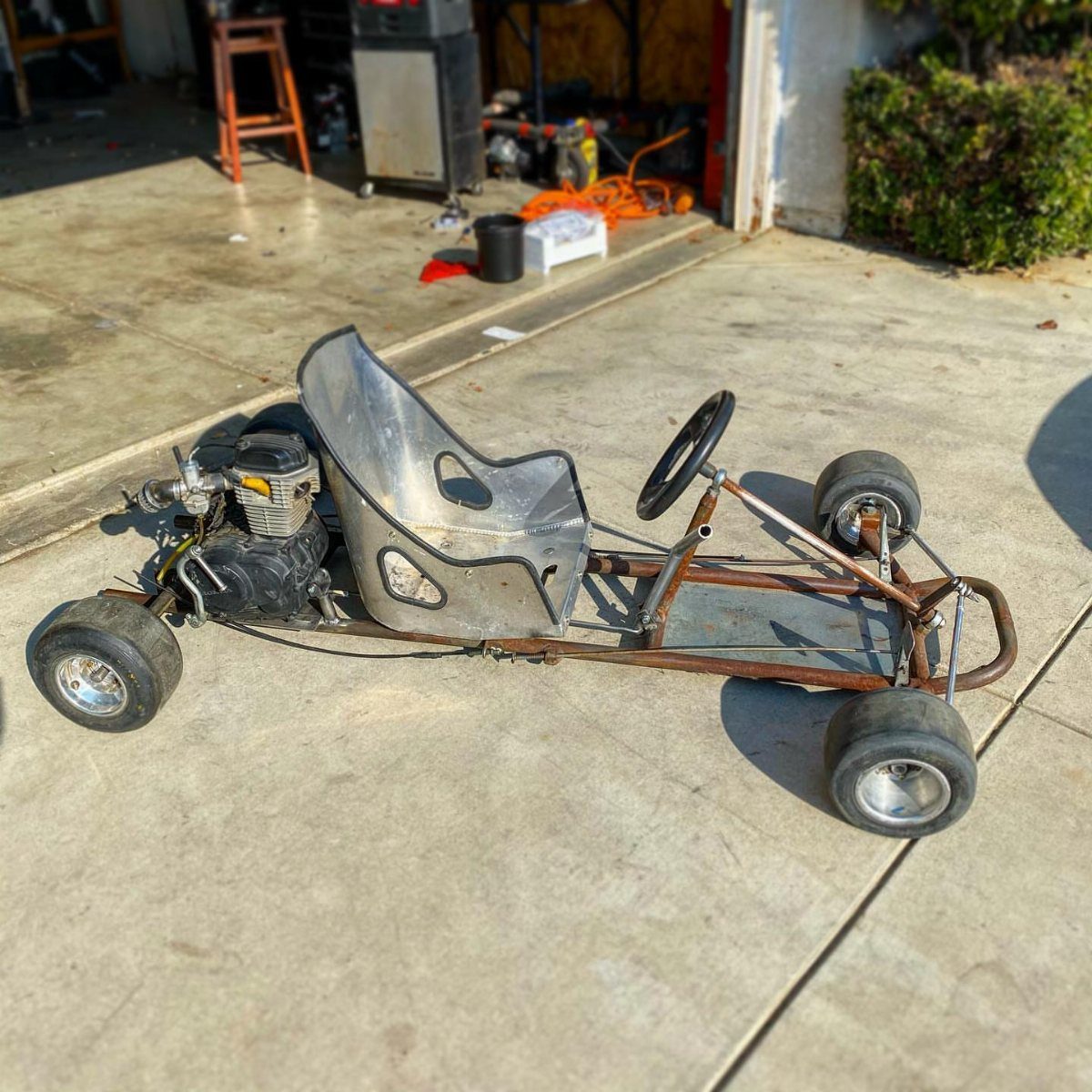 Homemade go-kart with a metal frame and silver seat on a concrete driveway.