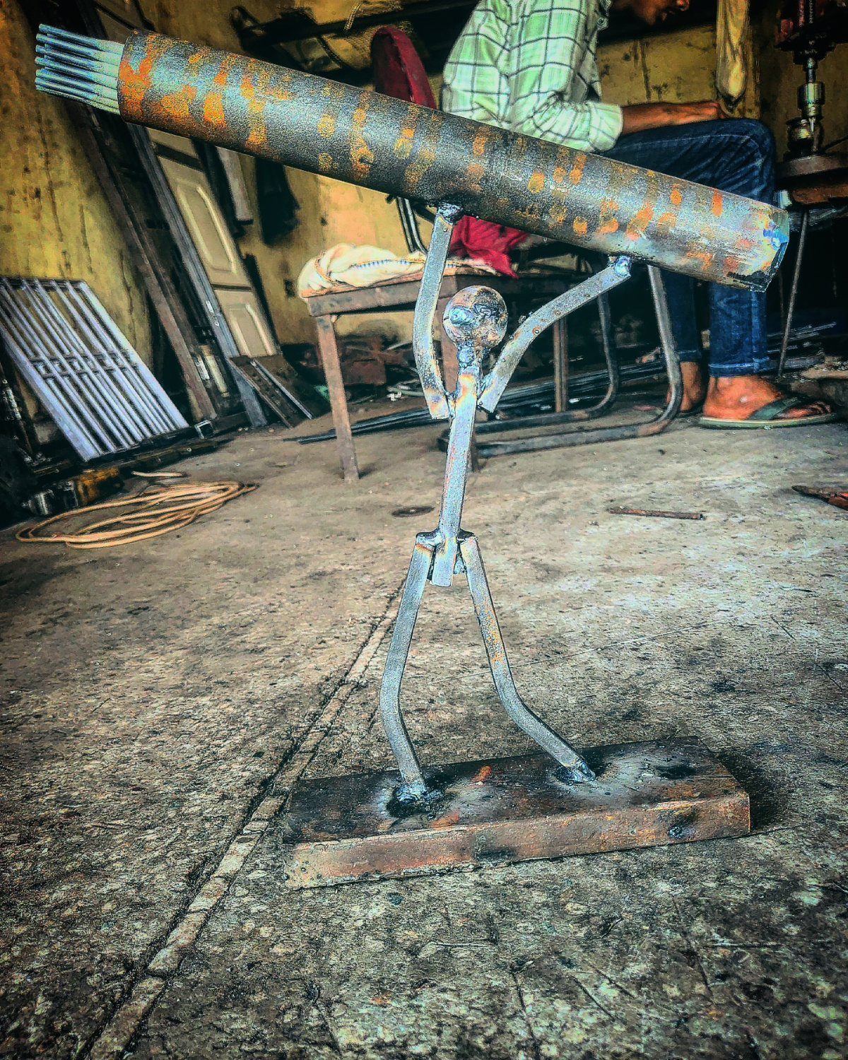 Welded metal stick figure sculpture holding a heavy rusted cylinder on its shoulders.