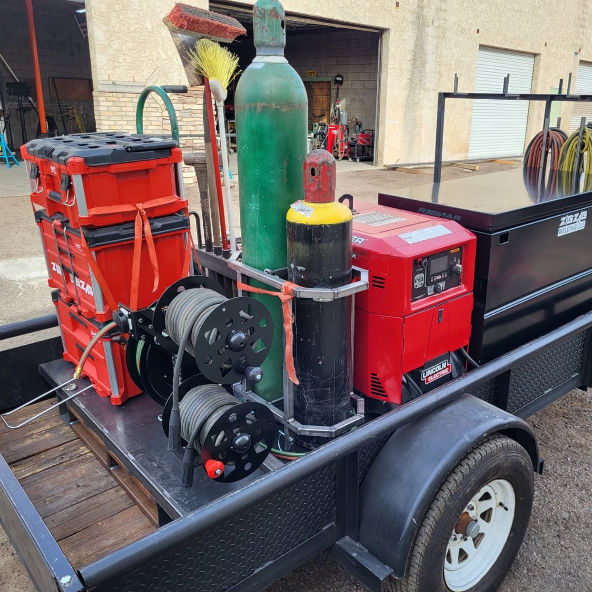 Black utility trailer equipped with a red welder, gas tanks, and tool chest.