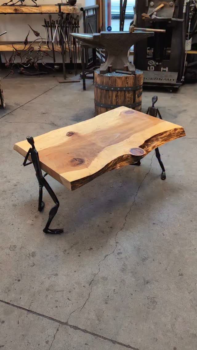 Live-edge wooden table with black metal legs shaped like human figures in a workshop.