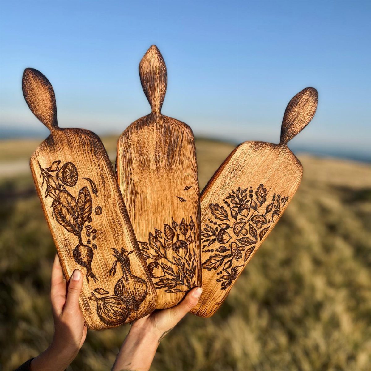 Three wooden cutting boards with detailed botanical wood burning art held outdoors.