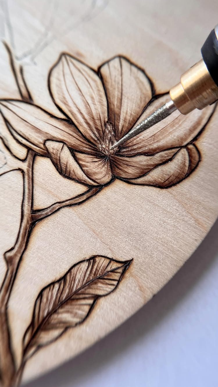 Wood burning pen creating a detailed magnolia flower with shaded petals on light wood.