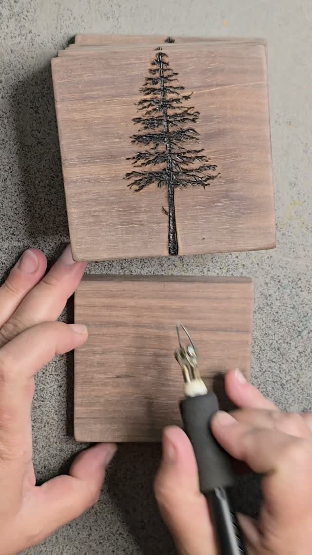 Hands using a wood burning tool on a square wooden coaster with a pine tree design.