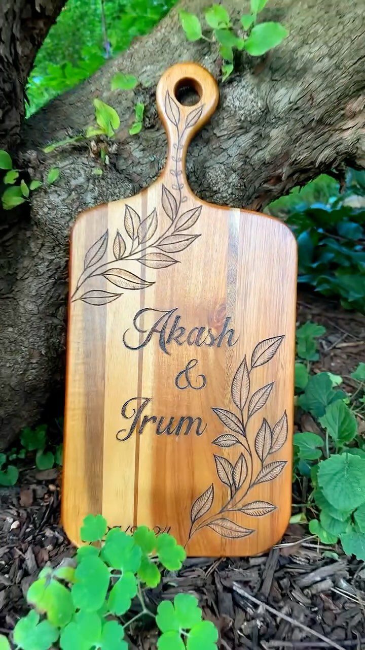 Wooden cutting board with wood-burned names and leaf designs leaning against a tree.