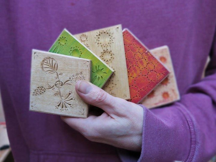 Hand holding five square wooden coasters with various carved geometric and floral patterns.