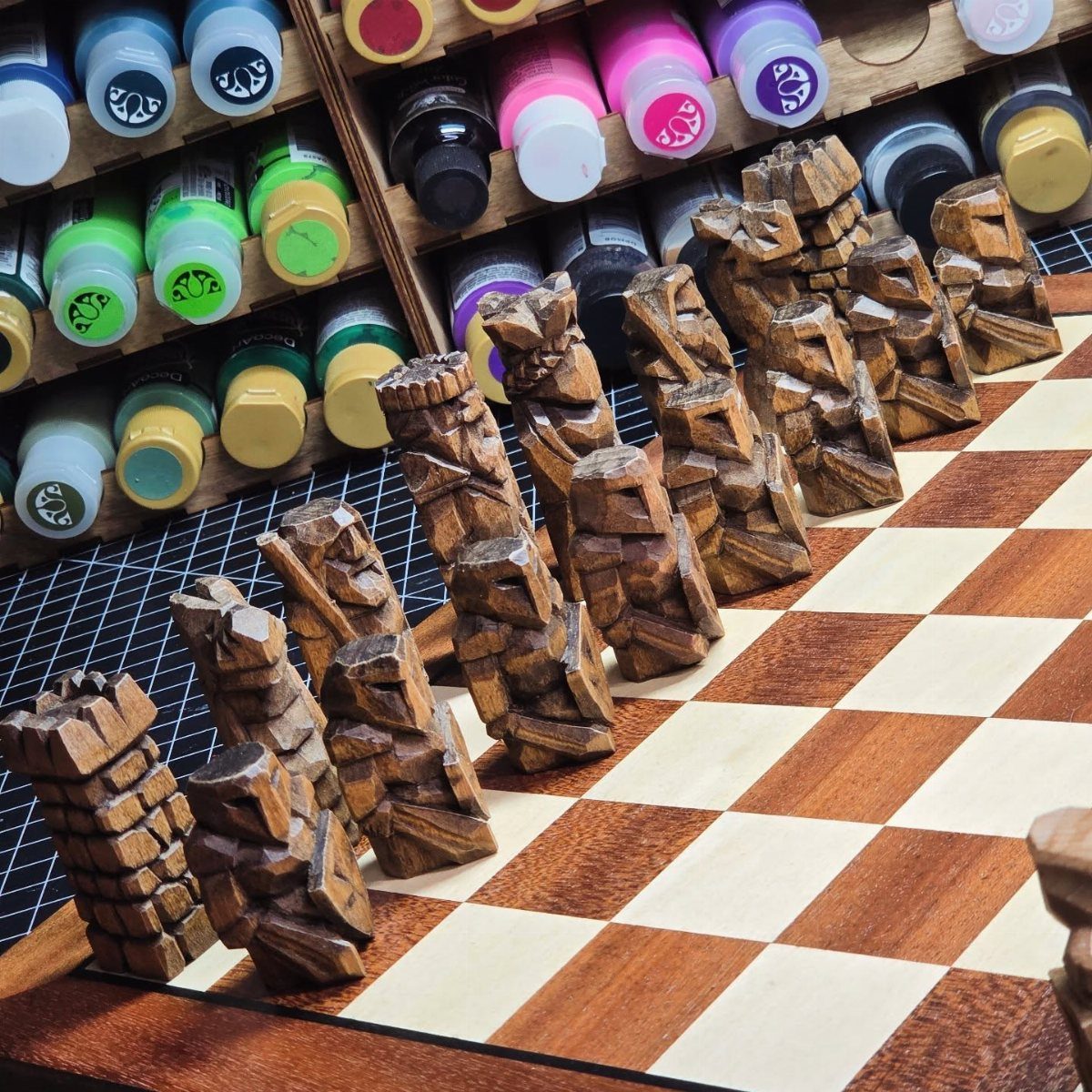 Tiki style hand-carved wooden chess pieces on a checkered wooden chessboard in a workshop.