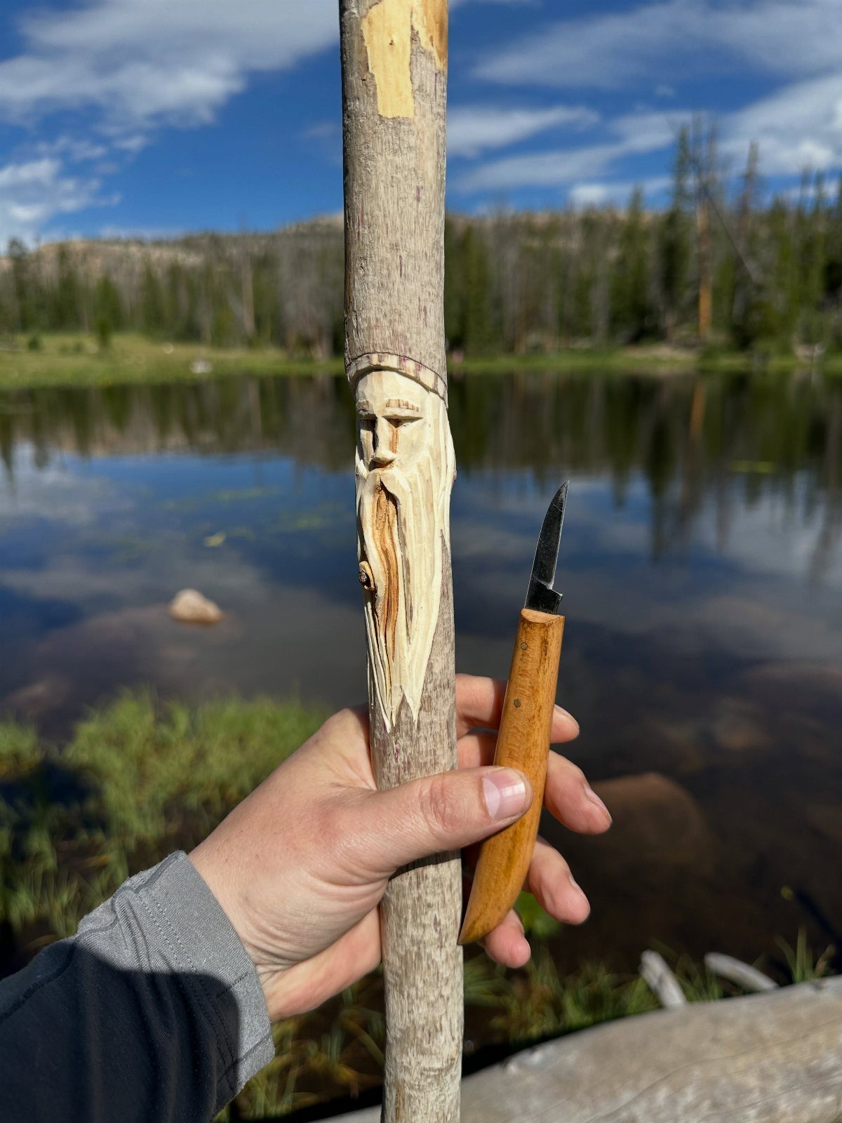Carved face on a wooden walking stick held next to a carving knife.