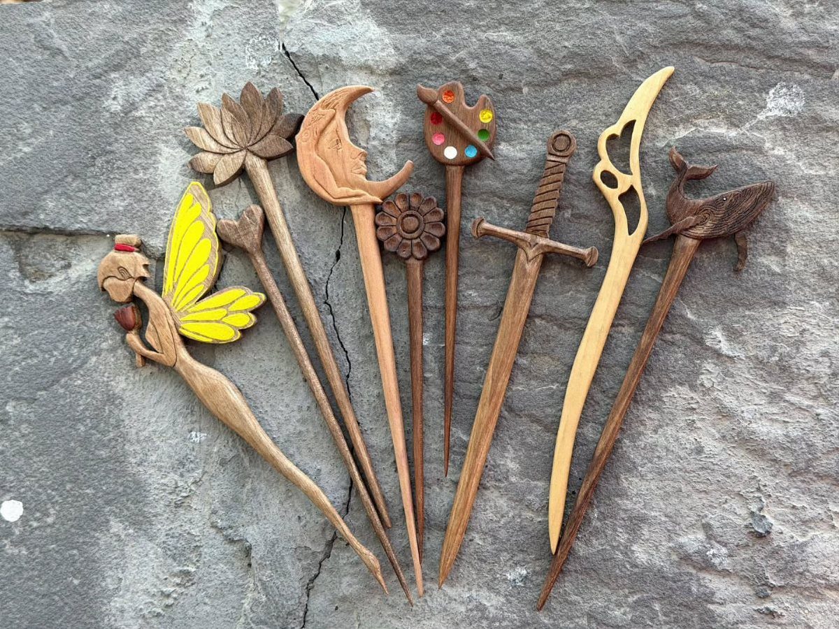 Assorted hand-carved wooden hair pins with decorative figures resting on a grey stone.