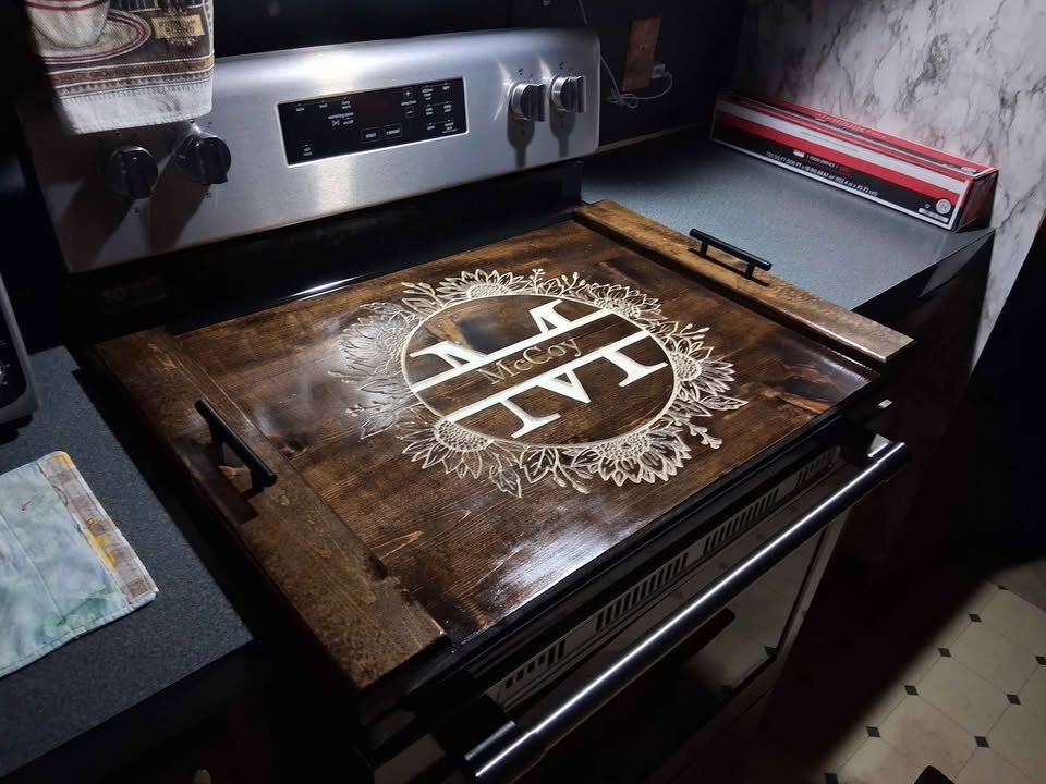 Wooden stove cover with engraved monogram resting on a stainless steel kitchen stove.