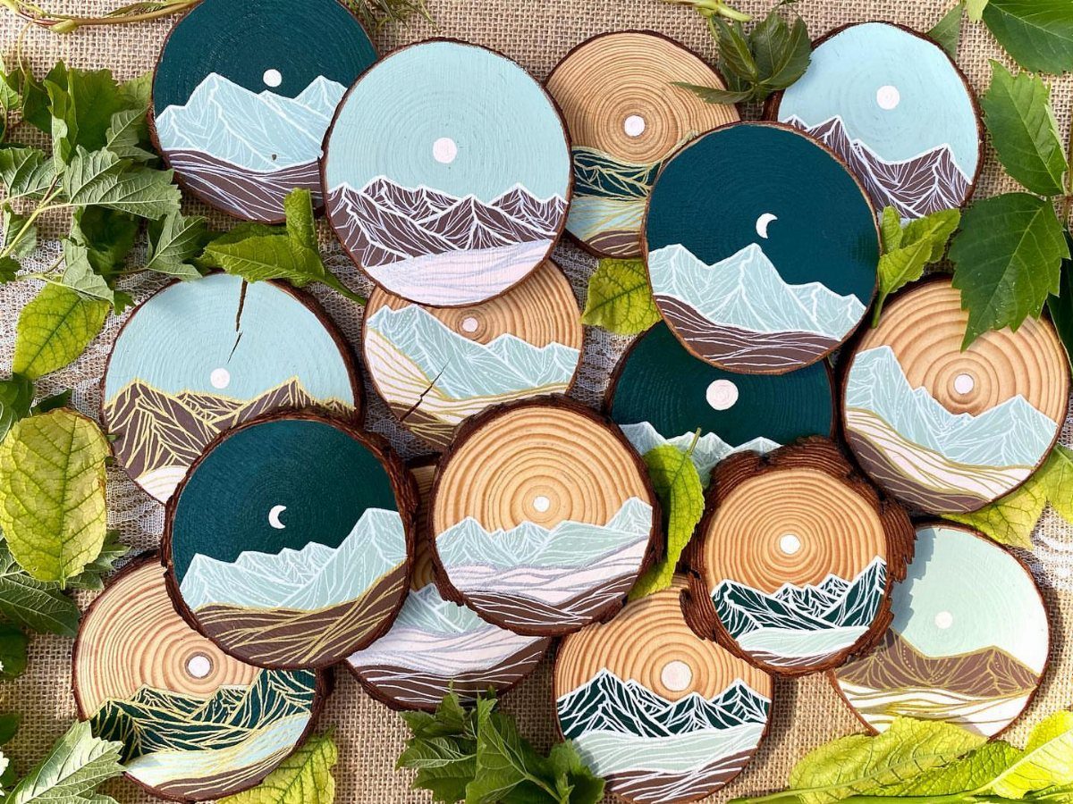 Circular wooden coasters painted with mountain landscapes resting on burlap with green leaves.