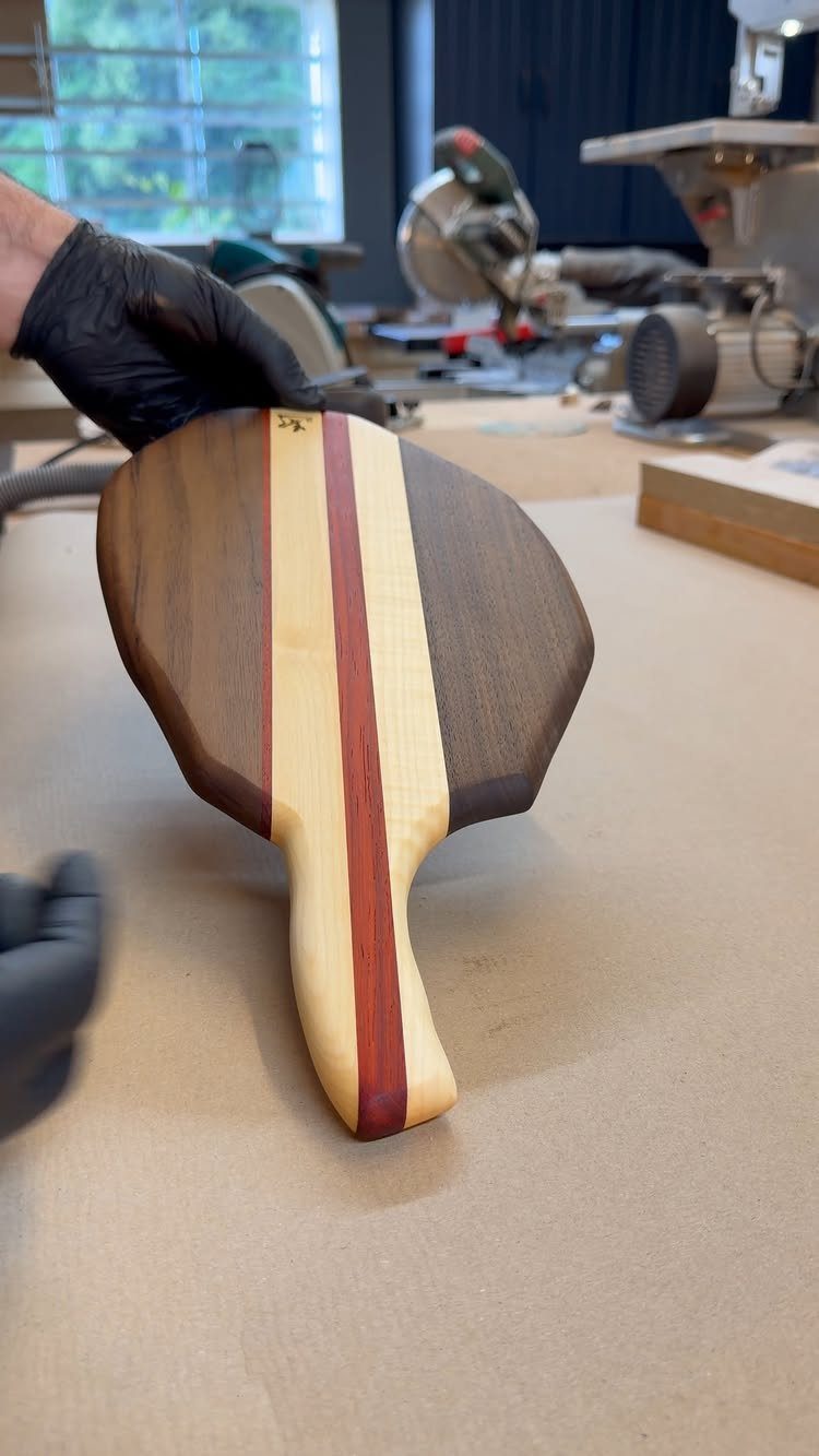 Handcrafted wooden cutting board with multi-colored wood stripes and a handle in a workshop.