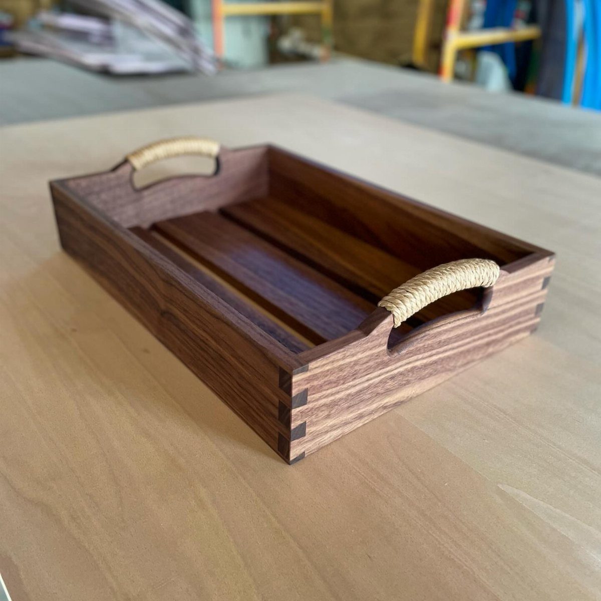 Dark brown wooden serving tray with rope handles on a light wood table.
