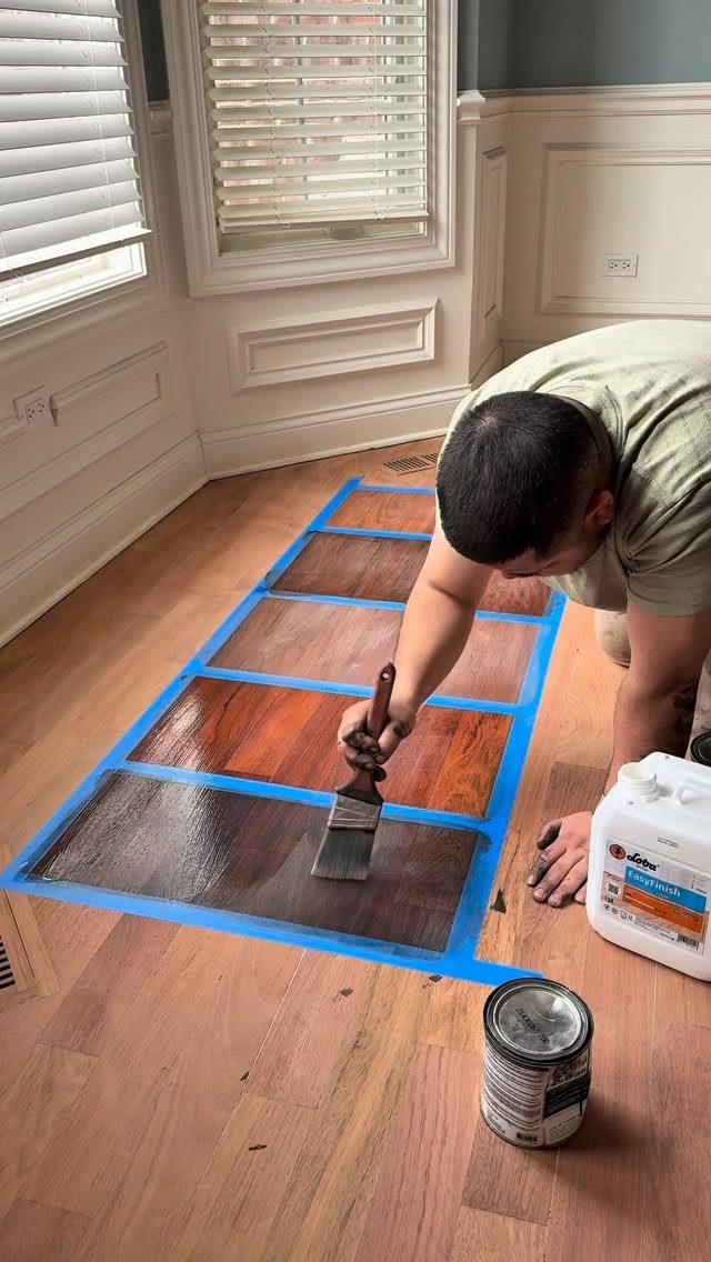 Man applying dark wood stain to a taped grid of floor samples.