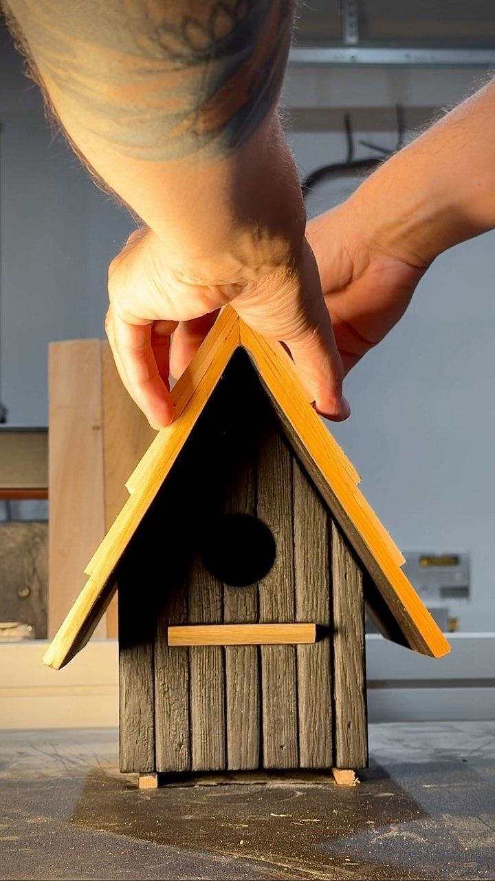 Hands assembling a dark grey wooden birdhouse with a bright yellow roof.