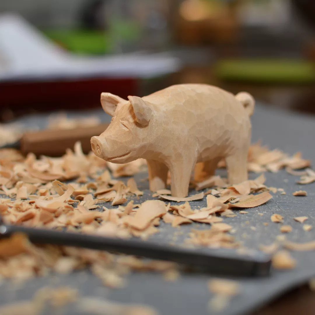 Hand-carved wooden pig figurine surrounded by wood shavings on a grey workbench