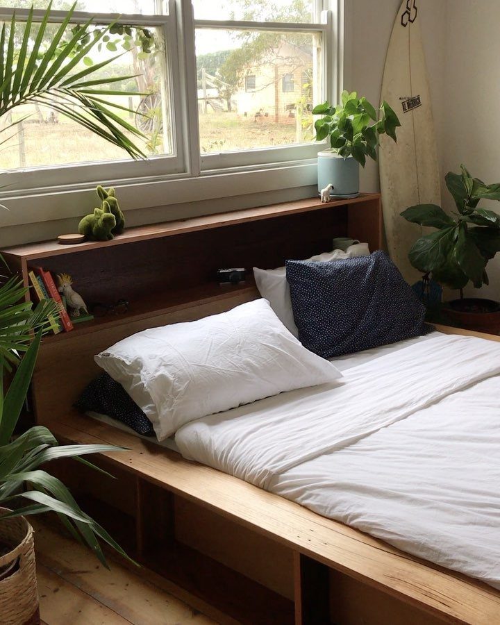 Wooden bed frame with built-in shelving, white bedding, and indoor plants by a window.