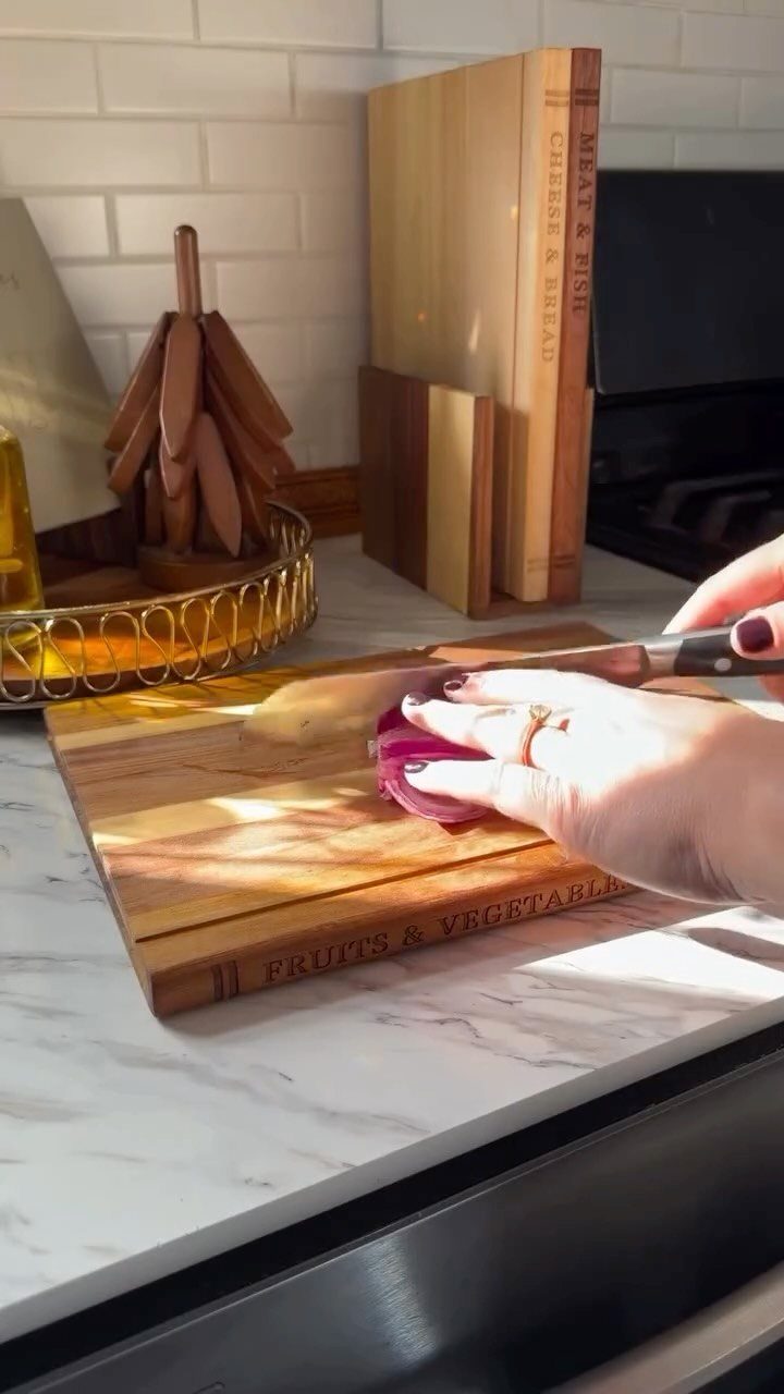 Hands slicing red onion on a wooden board with other boards standing behind.